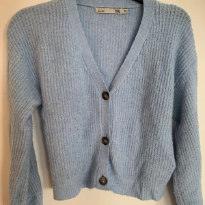 Ljusblå stickad kofta med knappar - Supermysig ljusblå stickad kofta med v-ringning och tre stora knappar framtill. Koftan har lång ärm och ribbad struktur som ger en chill och trendig look. Perfekt att slänga på sig när det är lite kyligare ute.