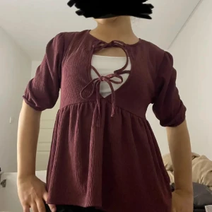 Vinröd blus med snörning - Säljer en vinröd blus med snygg snörning framtill och rundad hals. Blusen har puffiga, korta ärmar och ett flowigt fall nedtill 💕