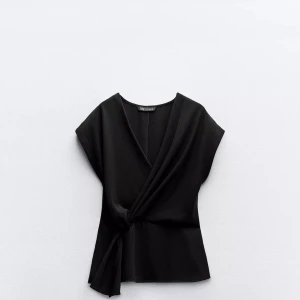 Svart omlottopp från Zara - Svart omlottopp från Zara med v-ringning och knyt i midjan. Toppen har korta ärmar och en stilren, elegant look. Materialet är mjukt och har ett lätt fall. Perfekt att styla med jeans eller kjol för en cool vibe.