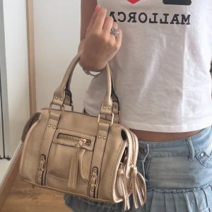 Beige handväska i skinn med detaljer - Snygg beige handväska i skinn med guldiga metalldetaljer, dragkedja framtill och dekorativa spännen. Väskan har två handtag och tofsdetaljer på sidorna för en trendig look. 
