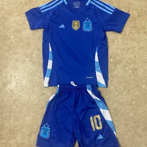 Argentina Messi blå fotbollströja Adidas - Säljer en blå Argentina fotbollströja med matchande shorts från Adidas. Tröjan har korta ärmar, guldtryck med nummer 10 och Messi på ryggen, samt AFA-emblem och FIFA-märke på bröstet. Materialet är lätt polyester med vita och ljusblå detaljer längs sidorna.