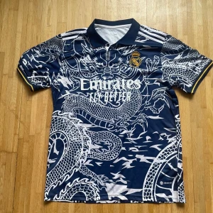 Real Madrid drakmönstrad fotbollströja - Snygg Real Madrid fotbollströja från Adidas med ett fett drakmönster i vitt på marinblå bakgrund. Tröjan har korta ärmar, krage och gula detaljer vid ärmslut. Materialet är lätt och andas, perfekt för match eller träning. Officiell klubb-logga på bröstet.
