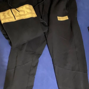 Svarta mjukisbyxor från BOSS - Svarta mjukisbyxor från BOSS med guldfärgad logga på benet. Byxorna har en avslappnad passform och är tillverkade i mjukt bomullsmaterial. Perfekta för en chill och stilren look.