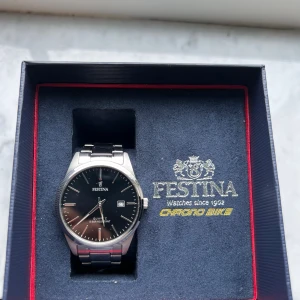 Festina herrklocka med svart urtavla - Stilren herrklocka från Festina med svart urtavla och datumvisning. Klockan har ett klassiskt runt silverfärgat boett och matchande länkarmband i metall. Perfekt accessoar för dig som gillar en tidlös och elegant look. Klockan har några små repor på undersidan men inget man ser jätte mycket.