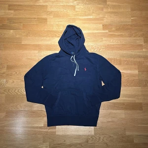 Marinblå hoodie från Polo Ralph Lauren - Snygg marinblå hoodie från Polo Ralph Lauren med klassisk röd logga på bröstet. Tröjan har huva med vit snörning och ficka. Skick 8/10