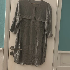 Grå skjortklänning med fickor - En grå skjortklänning med trekvartsärm och två stora bröstfickor. Klänningen har en rak passform och knäppning framtill. Perfekt för en avslappnad och stilren look. Vet ej storlek men gissar på M/L