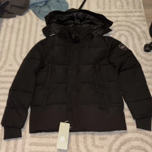 Svart dunjacka från Canada Goose - Säljer en svart dunjacka från Canada Goose med huva och klassisk patch på ärmen. Jackan har quiltad design, dragkedja och knappar framtill samt ribbade muddar vid ärmsluten. Perfekt för kalla dagar och riktigt snygg streetstil.