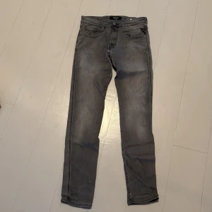 Replay Anbass grå jeans, storlek 31x32 - Snygga Replay Anbass jeans i tvättad grå färg med klassisk femficksdesign och svarta detaljer på bakfickorna. Slimmad passform och raka ben, tillverkade i mjukt jeanstyg med stretch för extra komfort. Perfekt för dig som gillar stilrena och moderna jeans.