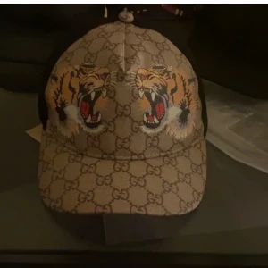 Gucci keps med tigertryck och GG-mönster - Säljer en snygg Gucci keps i beige med klassiskt GG-mönster och två stora tigerhuvuden framtill. Kepsen har svart mesh på sidorna och bak, samt justerbar rem. Märkt med Gucci både invändigt och utvändigt. Perfekt statement piece för dig som gillar lyxiga accessoarer.