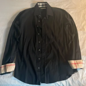 Svart Burberry skjorta med rutiga detaljer - Svart skjorta från Burberry i storlek S med klassiska rutiga detaljer på insidan av krage. Skicket är helt nytt! Skjortan har knappar framtill, lång ärm och en diskret broderad Burberry-logga på bröstet. Materialet är mjuk bomull med lite stretch för extra komfort. Lägg gärna prisförslag! 