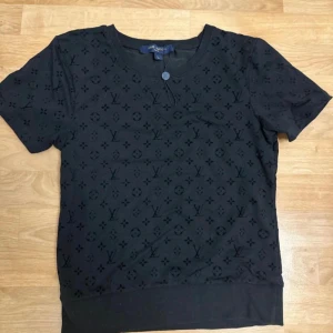 Svart Louis Vuitton t-shirt med monogram - Svart t-shirt från Louis Vuitton med klassiskt LV-monogram över hela tröjan. Rund halsringning, korta ärmar och mjukt material som känns skönt mot huden. Perfekt för dig som vill ha en stilren och lyxig look med tydlig logga.