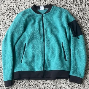 Turkos fleecejacka från Wear Colour - Snabb leverans.  Turkos fleecejacka från Wear Colour med svarta detaljer vid muddar, krage och fickor. Jackan har hel dragkedja, två sidofickor med dragkedja och en extra ficka på ärmen. Mjuk och skön med sportig vibe, perfekt för kyliga dagar.