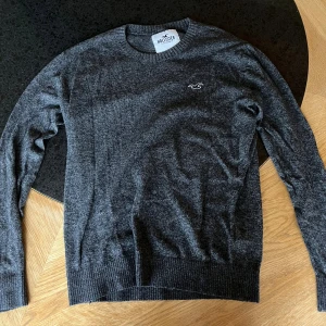 Mörkgrå stickad tröja från Hollister - Snygg mörkgrå stickad tröja från Hollister med diskret logga på bröstet. Tröjan har rund hals, ribbade muddar och lång ärm. Perfekt för dig som gillar enkel och stilren look. Passar dig som vill ha en normal passform och mjukt material.