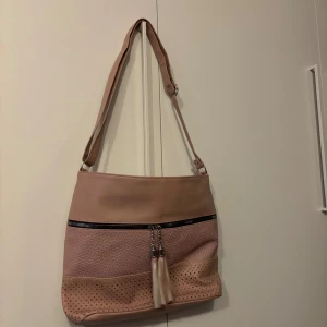 Rosa axelväska med tofsdetalj - Snygg rosa axelväska med justerbar rem och coola tofsar vid dragkedjan. Väskan har en mix av slät och perforerad yta i syntetmaterial, vilket ger en modern vibe. Perfekt storlek för att få plats med allt du behöver och passar till många olika stilar.