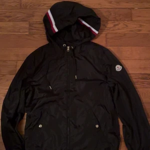 Svart vindjacka från Moncler med huva - Svart vindjacka från Moncler med huva som har röd och vit rand. Jackan har dragkedja framtill, två fickor med tryckknappar och Moncler-logga på ärmen. Tillverkad i glansig polyester och har snygga detaljer som snörning i huvan.