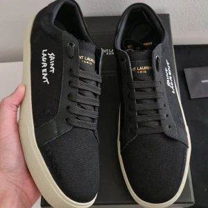 Svarta Saint Laurent sneakers - Svarta sneakers från Saint Laurent med vit logotyp på sidan och guldtext på innersulan. Skorna har snörning, rund tå och en tjock, vit platt sula med fiskbensmönster undertill. Ovandelen är i textil och skinn, vilket ger en lyxig och stilren vibe. Riktigt feta skor till höst/vinter. Storlek 43! 💥 Box och saint laurent dustbag medföljer! Pris kan diskuteras vid snabb affär.