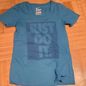Blå Nike Dri-Fit t-shirt Just Do It - Blå Nike Dri-Fit t-shirt med trycket 'JUST DO IT' i mörkblått framtill. Klassisk rund halsringning och korta ärmar. Tillverkad i ett lätt och snabbtorkande syntetmaterial som passar perfekt för träning. Snygg och sportig stil med liten Nike-logga vid nederkanten.