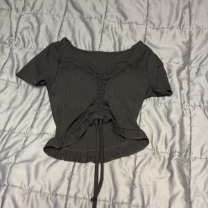 Svart croppad topp med snörning - Snygg svart croppad topp med korta ärmar och ribbad struktur. Toppen har en dragsko med snörning framtill som ger en cool rynkad effekt och gör att du kan justera passformen.