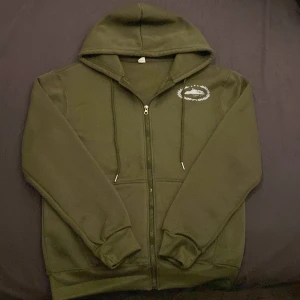 Olivgrön Corteiz hoodie med dragkedja - Säljer en olivgrön hoodie från Corteiz med stor vit logga på ryggen och mindre tryck på bröstet. Tröjan har huva med snörning, dragkedja framtill och två fickor. Mjuk och skön, perfekt för chill dagar.