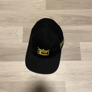 Svart keps från Speed med gul brodyr - Svart keps från Speed med justerbar snapback och gul broderad logga framtill. Kepsen har böjd skärm och detaljer i gult på sidan. Tillverkad i polyester och bomull för en bekväm passform.