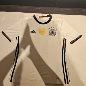 Tysklands VM 2014 fotbollströja Adidas - Säljer en officiell Tysklands landslagströja från VM 2014 med FIFA World Champions-märke. Tröjan är vit med svarta detaljer, svarta ränder längs sidorna och svart krage. Korta ärmar med röd, svart och gul kant. Materialet är lätt och andas, climacool-teknologi.