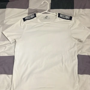 Vit Moschino t-shirt med logga - Vit t-shirt från Moschino med svart logga på axlarna och i nacken. Klassisk rund halsringning och korta ärmar. Tillverkad i mjuk bomull för en skön känsla. Enkel men ändå stilren design som sticker ut med de tydliga loggdetaljerna.