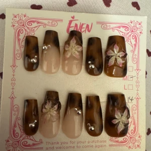 Två set med dekorerade lösnaglar - Säljer två set lösnaglar. Första setet har bruna och nude toner med handmålade blommor och silverfärgade stenar, i oval form. Andra setet är spetsiga med rosa och vita detaljer, handmålade rosetter och blommor samt guldiga pärlor. Perfekt för dig som vill ha unika naglar med 3D-effekt.