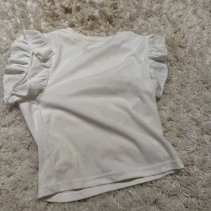 Vit topp med volangärmar - Säljer en söt vit topp med korta volangärmar. Toppen är ribbad och har en rak passform som känns både trendig och enkel att styla. Perfekt för dig som gillar en feminin touch på din outfit.