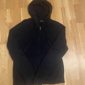 Stickad marinblå zip up hoodie från Lexington - Marinblå hoodie från Lexington med dragkedja framtill och huva med snörning. Tröjan har långa ärmar och är tillverkad i mjukt bomullsmaterial. Perfekt för en chill och avslappnad stil.