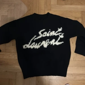 Svart Saint Laurent stickad tröja M - Svart stickad tröja från Saint Laurent med vit, handskriven logga framtill. Tröjan har rund hals och långa ärmar, tillverkad i Italien. Perfekt för dig som gillar exklusiva och snygga designerplagg med statement-tryck.