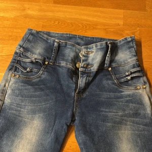 Blå jeans med snörning från Top M Jeans - Snygga blå jeans med cool snörningsdetalj både fram och bak, samt guldfärgade nitar vid fickorna. Modellen är högmidjad och har en slim passform med raka ben. Jeansen har en tvättad look och är tillverkade i klassiskt denimtyg.