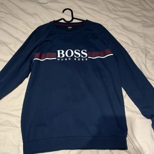 Marinblå sweatshirt från Hugo Boss - Tja säljer en Snygg marinblå sweatshirt från Hugo Boss med vit och vinröd rand över bröstet och stor BOSS-logga. Tröjan har långärmad passform och rund hals. Storlek L 