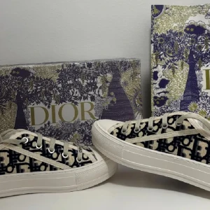 Dior sneakers med monogram och snörning - Snygga Dior sneakers med klassiskt monogrammönster i svart och beige. Skorna har rund tå, platt vit sula med svart stjärndetalj undertill och snörning framtill. Ovandelen är i textil med tydligt Christian Dior-tryck på plösen. Perfekt för dig som gillar lyxiga streetwear-vibbar.