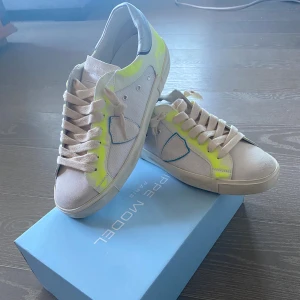 Philippe Model sneakers med neon-detaljer - Hej säljer minna snygga sneakers från Philippe Model i vitt med neon-gula detaljer och blå kant runt loggan på sidan. Skorna har rund tå, platt sula och breda snören. Ovandelen är i en mix av skinn och textil, med grå hälparti och coola kontraster. Perfekt för dig som gillar att sticka ut. Hör gärna av dig om du är intresserad 