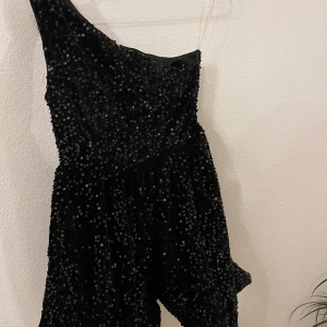 Svart glittrig oneshoulder klänning XS - Supersnygg svart kortklänning från Faeriesty med glittriga paljetter över hela och en cool oneshoulder-design. Perfekt för dig som vill sticka ut med en unik look. Klänningen har en figurnära passform och är riktigt festlig.
