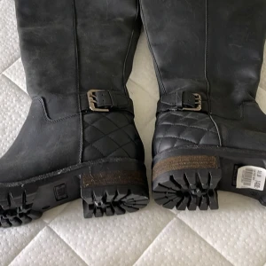 Svarta quiltade boots med spänne  - Snygga svarta boots med quiltad detalj och spänne vid ankeln. Grov sula för extra grepp och cool look. Tillverkade i skinn med robust känsla och rund tå. Perfekta för dig som vill ha en edgy stil med lite biker-vibe. Från scorett 