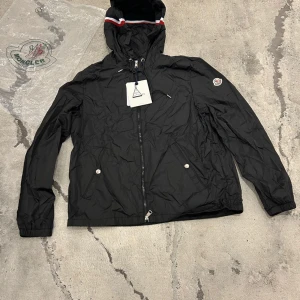 Svart vindjacka från Moncler - Svart vindjacka från Moncler med huva och dragsko, klassisk logga på ärmen och snygga detaljer i rött och vitt på huvan. Jackan har dragkedja framtill, två fickor med tryckknappar och är tillverkad i lätt polyester. Perfekt för dig som vill ha en stilren och sportig look.