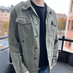 Grön militär overshirt - Säljer en grön militärinspirerad overshirt med tyska flaggan på ärmen. Jackan har två stora bröstfickor med lock och knappar. Jackan har avslappnad passform, perfekt att slänga över en hoodie eller t-shirt eller bara lager på lager nu till hösten.