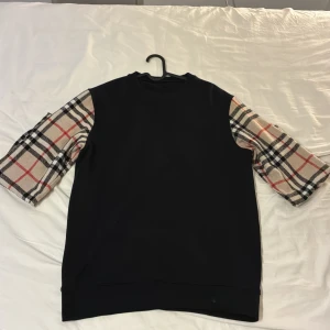 Svart sweatshirt med rutiga ärmar - Cool svart sweatshirt med klassiska beige-rutiga ärmar i Burberry-inspirerat mönster. Tröjan har rund hals och ribbad nederkant. Perfekt för dig som vill sticka ut med en unik stil och gillar kontraster mellan svart och mönstrat.