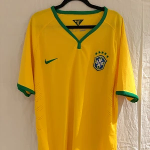 Brasilien Nike fotbollströja gul/grön - En retro Brasilien fotbollströja från Nike i storlek L. Tröjan är gul med gröna detaljer vid krage och ärmslut, har broderat brasilien märke med fem stjärnor på bröstet och Nike-logga. Tillverkad i lätt, ventilerande Dri-Fit polyester för maximal komfort.