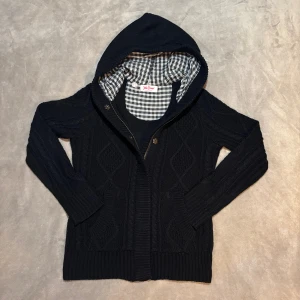 Svart stickad hoodie - Svart stickad hoodie med snyggt rutigt foder i huvan. Tröjan har dragkedja, knappar och ribbade muddar. Mönstrad med diskreta diamantformer och har två fickor framtill. Perfekt för dig som gillar chill och stil. 🦦 