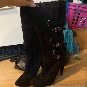 Svarta stövletter med klack och spännen - Snygga svarta stövletter med hög klack och flera dekorativa spännen i guld längs skaftet. Gjorda i mocka för en mjuk och lyxig känsla. Rund tå och dragkedja på insidan för enkel påtagning. Perfekt för dig som vill ha en edgy look.