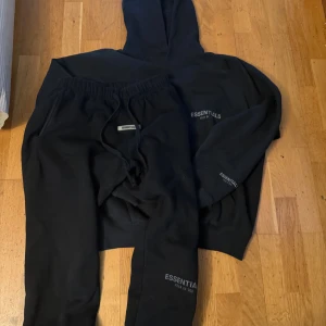 Svart Essentials hoodie från Fear of God - Svart hoodie från Fear of God Essentials med diskret logga på bröstet och ärmen. Tröjan har huva, känguruficka och långa ärmar. Mjuk och skön bomullskvalitet, perfekt för chill eller streetwear. Snygg och enkel design som funkar till allt.