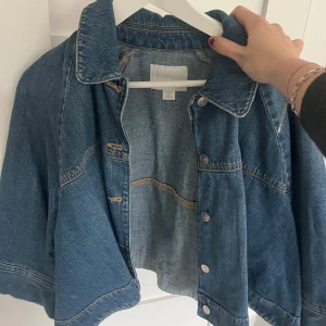 Blå croppad jeansjacka - Säljer en blå croppad jeansjacka med klassisk krage och knappar framtill. Jackan har vida trekvartsärmar med spänndetaljer vid ärmsluten och markerade sömmar för en cool look. Perfekt till vårens outfits!