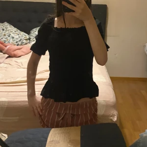 Svart topp med volang - Säljer en svart blus med korta puffärmar och söt volang nedtill. Blusen har en fyrkantig halsringning och är figurnära med en lätt ribbad struktur. Perfekt för dig som gillar en feminin och trendig stil.