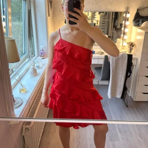 Röd one shoulder volangklänning ifrån Bubbleroom - Säljer en röd kortklänning med one shoulder och tunna axelband. Klänningen har flera lager volanger och är figurnära med dragkedja i sidan. Materialet är glansigt och känns lätt, perfekt för dig som vill sticka ut med en färgstark look. Storlek S. Aldrig använd med lappen kvar ❤️