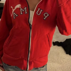 Röd hoodie från Puma med dragkedja - Röd hoodie från Puma med vit broderad logga och stora bokstäver framtill. Tröjan har huva med snörning, fickor på sidorna och hel dragkedja. Storlek m men sitter som s