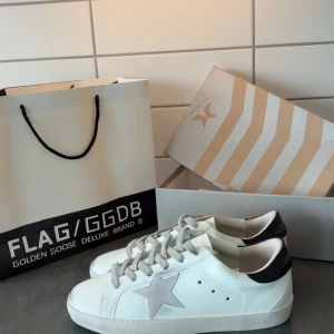 Golden Goose vita sneakers  - Golden Goose sneakers i vitt skinn med grå mockadetaljer och ikonisk stjärna på sidan. Skorna har grå snörning, svart hälparti med logga och platt sula. Perfekta för dig som gillar stilrena och trendiga sneakers med en lyxig touch.