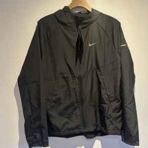Svart vindjacka från Nike med huva - Svart vindjacka från Nike med huva och dragkedja framtill. Jackan har en stilren design med diskret Nike-logga på bröstet och reflekterande detalj på ärmen. Perfekt för löprundan eller när du vill ha en sportig look.