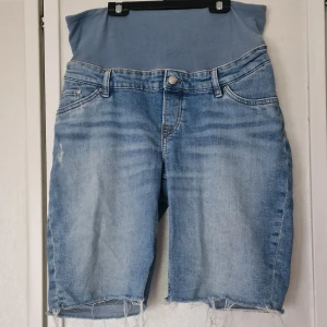 Blå denim mamma-shorts från H&M - Säljer ett par blå denim mamma-shorts från H&M med låg midja och mjuk ribbad magpanel. Klassisk femficksmodell, slitna detaljer och rå kant nedtill. Perfekta för dig som vill ha bekväma och snygga shorts under graviditeten.